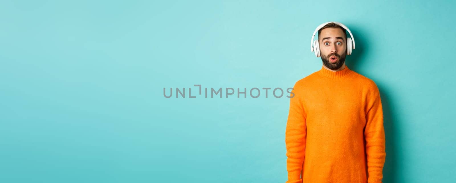 Amazed adult man listening music in headphones, looking at camera impressed with sound, standing over turquoise background.