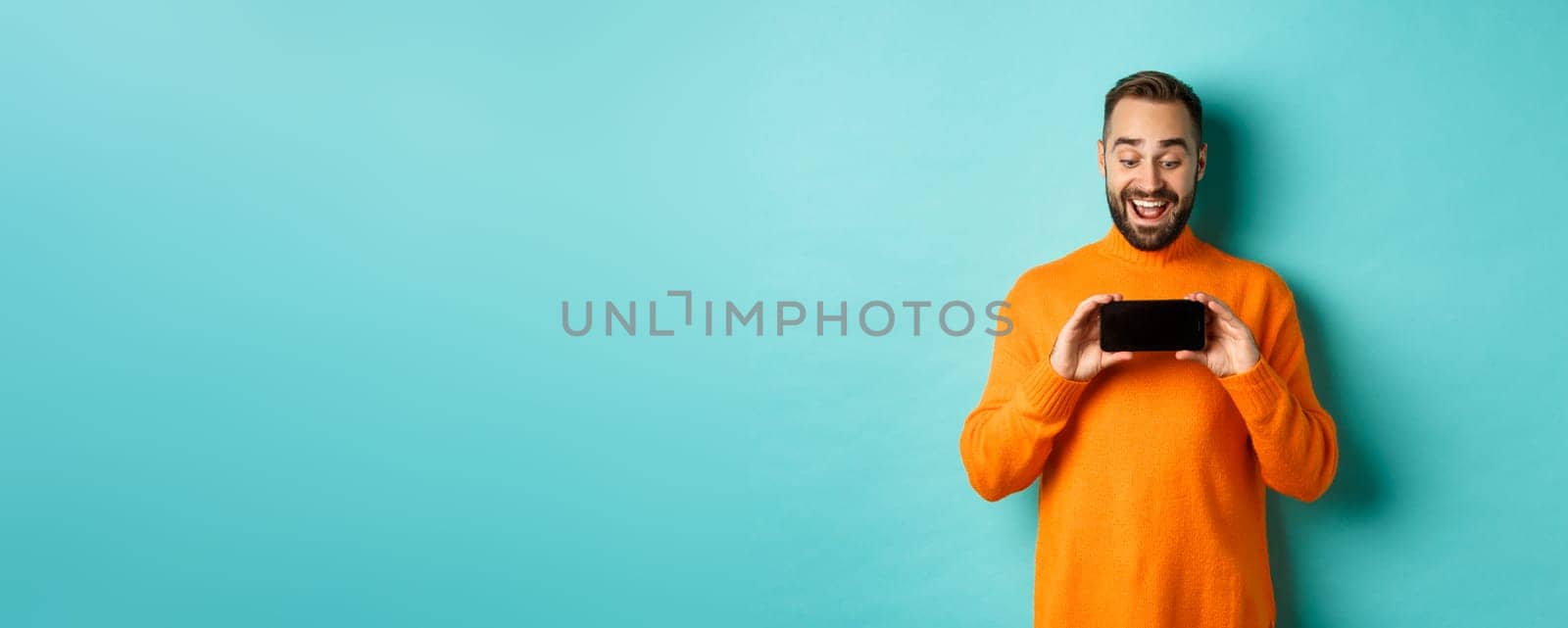 Excited guy demonstrating online store, showing mobile screen and looking amazed, standing over light blue background by Benzoix