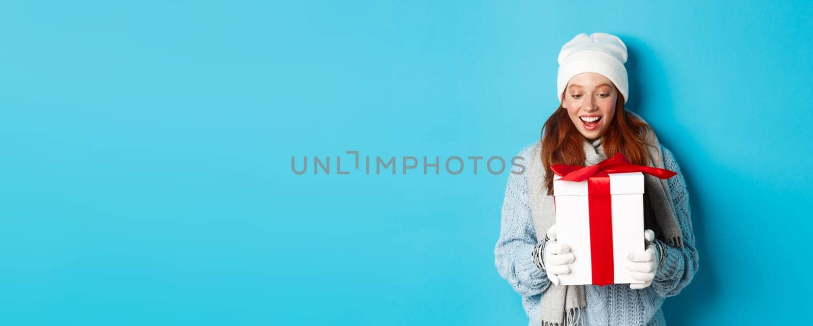 Winter holidays and Christmas eve concept. Surprised cute redhead girl in beanie and sweater receiving New Year gift, looking at present amazed, standing over blue background by Benzoix