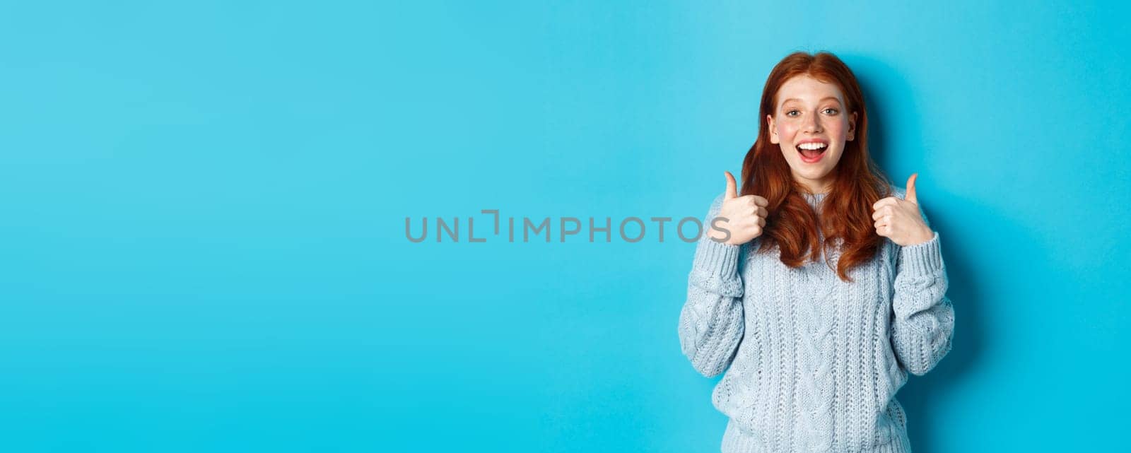 Supportive redhead girl in sweater, showing thumbs-up and looking amazed, praising good choice, standing over blue background.