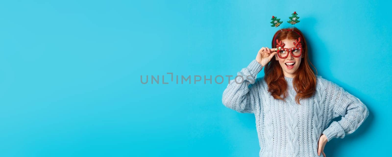 Christmas party and celebration concept. Cute redhead teen girl celebrating New Year, wearing xmas tree headband and funny glasses, looking left amused, blue background by Benzoix