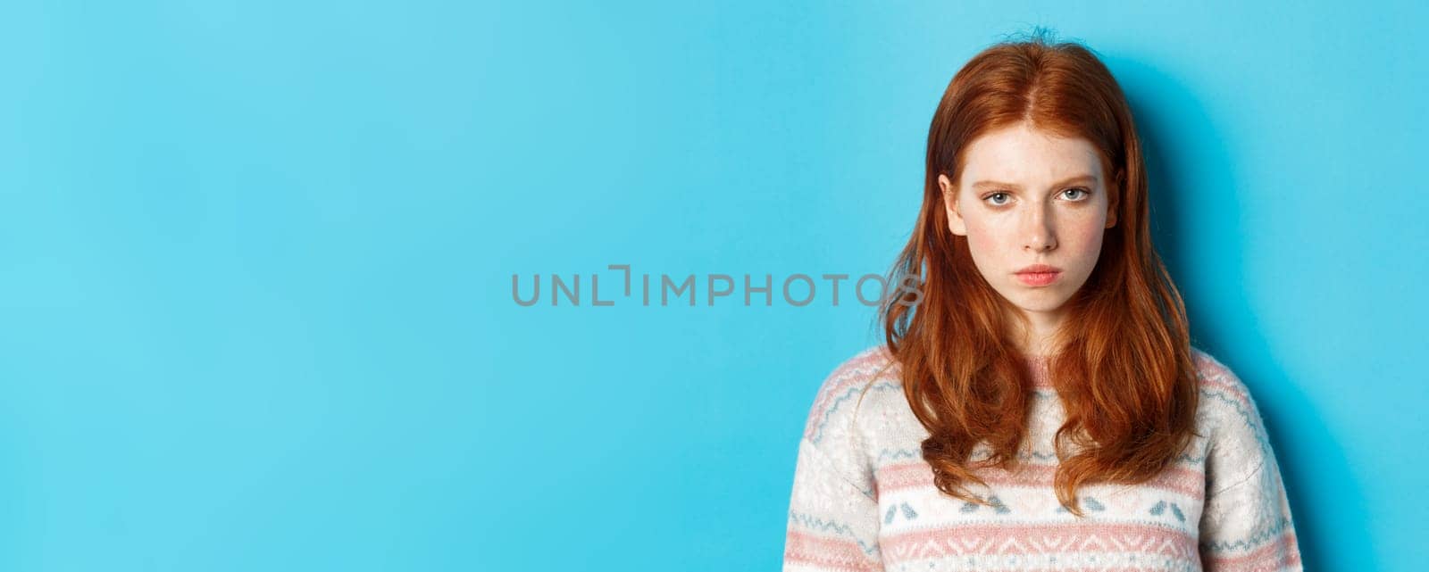 Close-up of angry redhead teenage girl looking pissed-off at camera, frowning and sulking mad, standing over blue background.