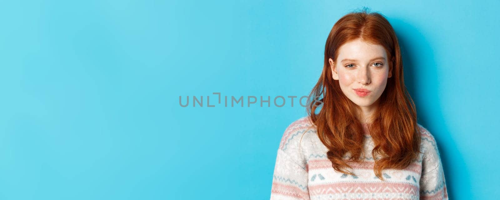 Close-up of redhead cunning girl having an idea, smirk and stare at camera satisfied with plan, standing over blue background.