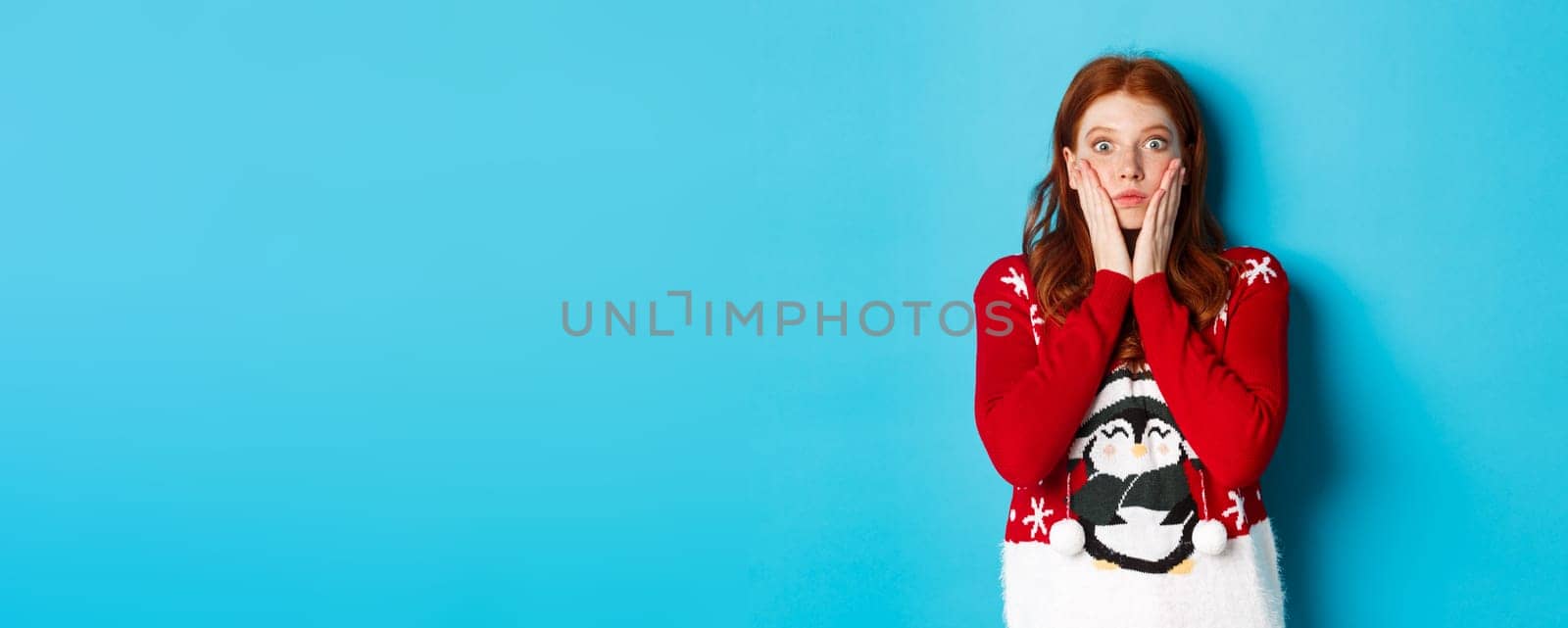 Winter holidays and Christmas Eve concept. Impressed and speechless redhead girl gasping, staring at camera with disbelief, standing in xmas sweater against blue background by Benzoix