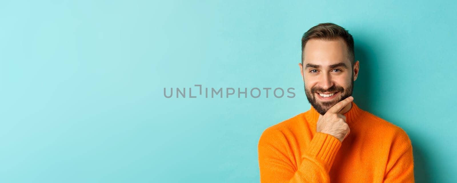 Close-up of handsome and confident man with beard smiling, looking thoughtful at camera, have plan, standing over light blue background.