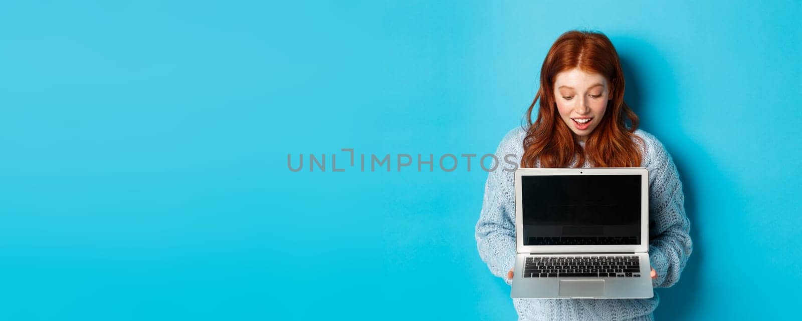 Amazed redhead girl staring at laptop screen and looking impressed, showing computer display, standing over blue background.