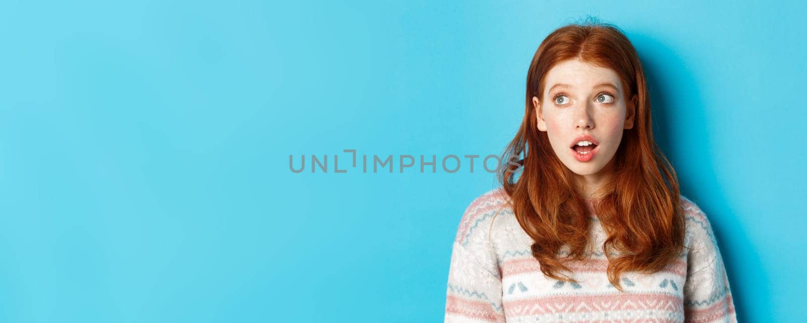 Close-up of impressed redhead girl staring left amazed, open mouth in awe and glancing at promo, standing in winter sweater against blue background by Benzoix