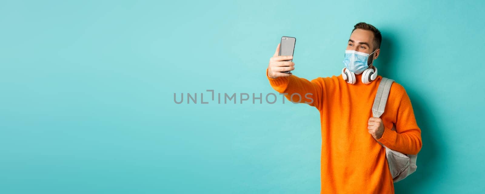 Young modern man with headphones and backpack, taking selfie on mobile phone in medical mask, standing over light blue background.