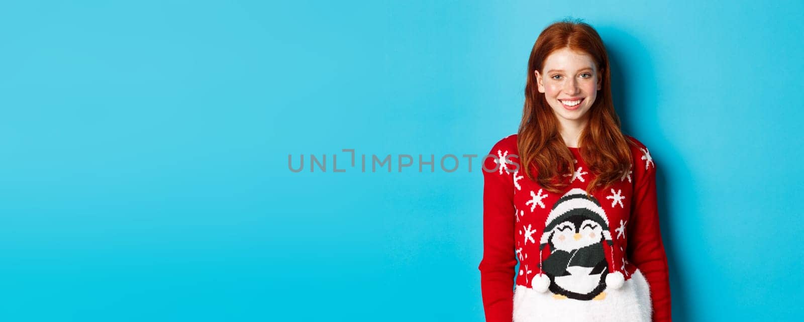 Winter holidays and Christmas Eve concept. Cute smiling teenage girl with red hair, wearing funny xmas sweater, standing against blue background.