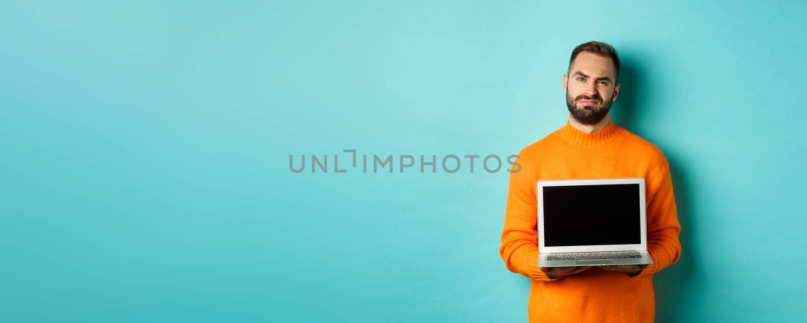 Handsome bearded man in orange sweater showing laptop screen, demonstrating promo, grimacing disappointed and upset standing over light blue background.