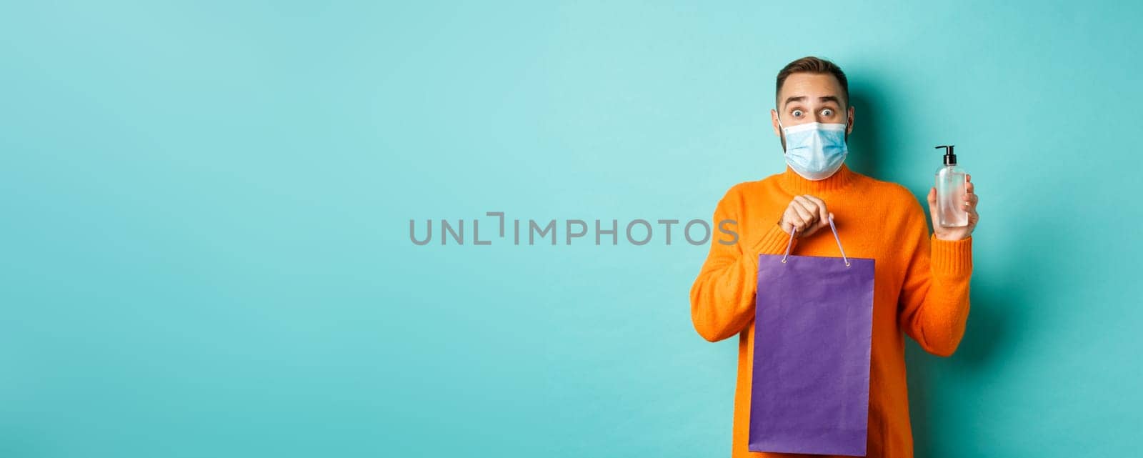 Coronavirus, pandemic and lifestyle concept. Man in face mask showing shopping bag and hand sanitizer, standing over turquoise background by Benzoix