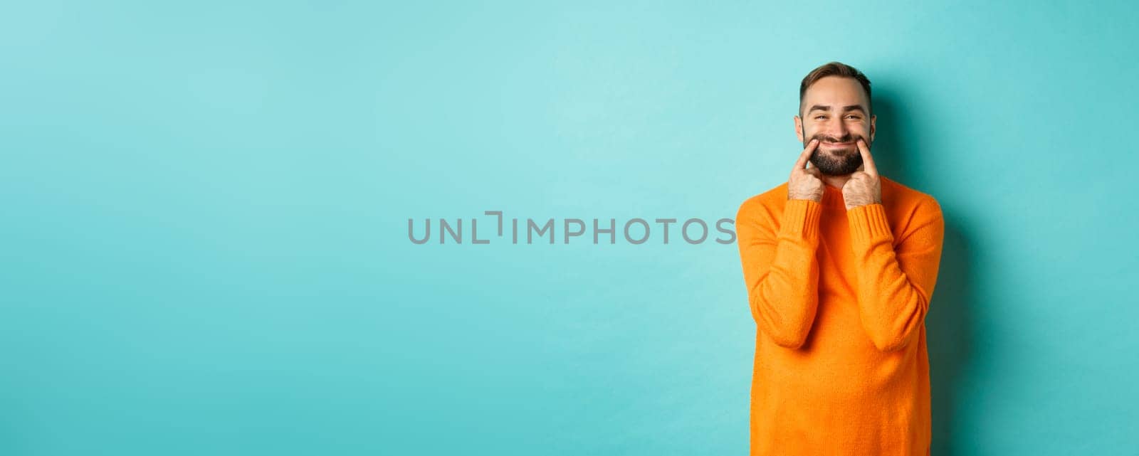 Image of bearded man stretching lips in happy smile, faking happiness, standing over light blue background. Copy space