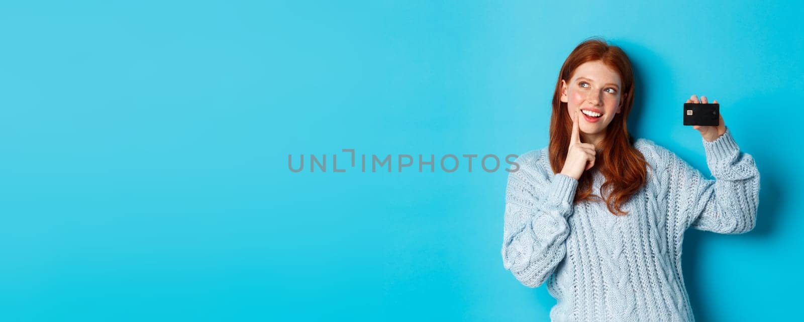 Image of thoughtful redhead girl thinking about shopping, showing credit card and pondering, standing over blue background by Benzoix