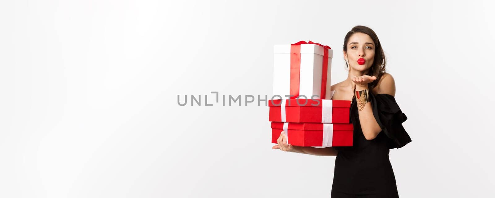 Celebration and christmas holidays concept. Pretty woman in elegant black dress holding presents, sending air kiss at camera, standing over white background by Benzoix