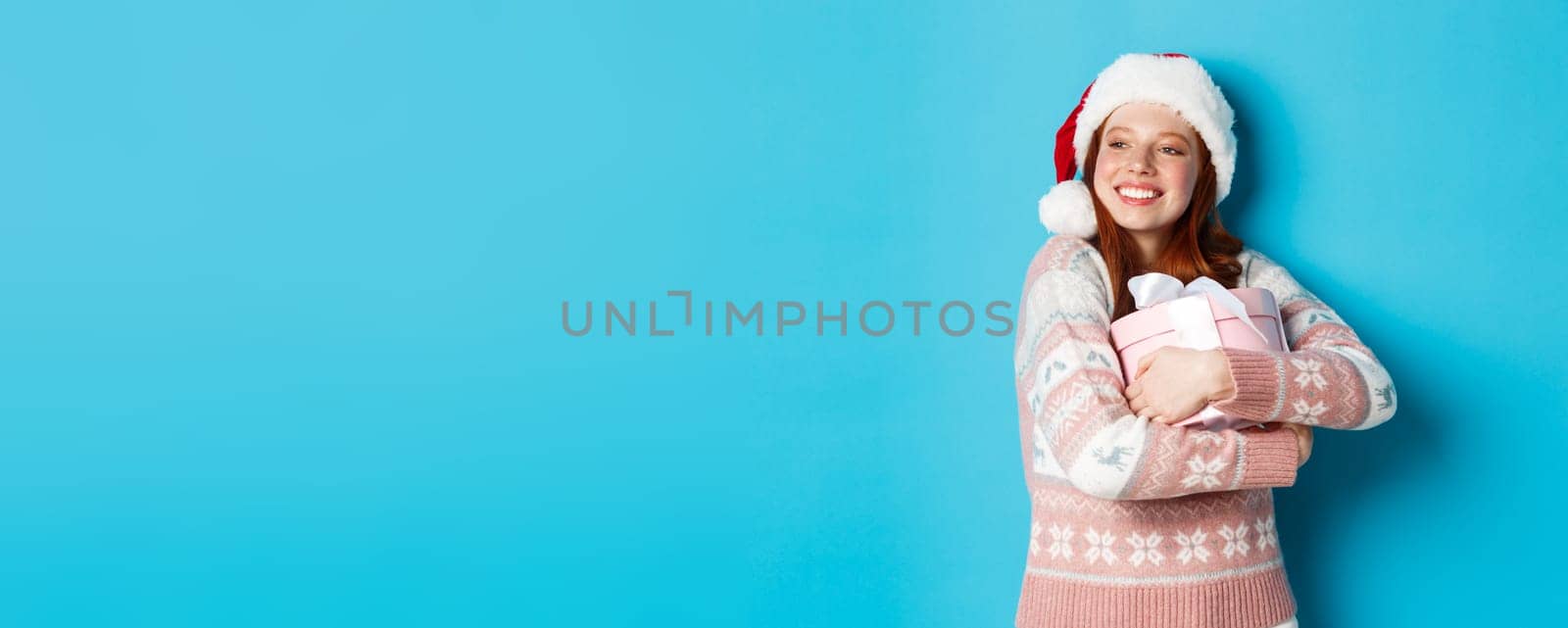 Winter and celebration concept. Dreamy redhead girl in santa hat hugging her christmas gift and looking left, smiling happy, standing over blue background by Benzoix