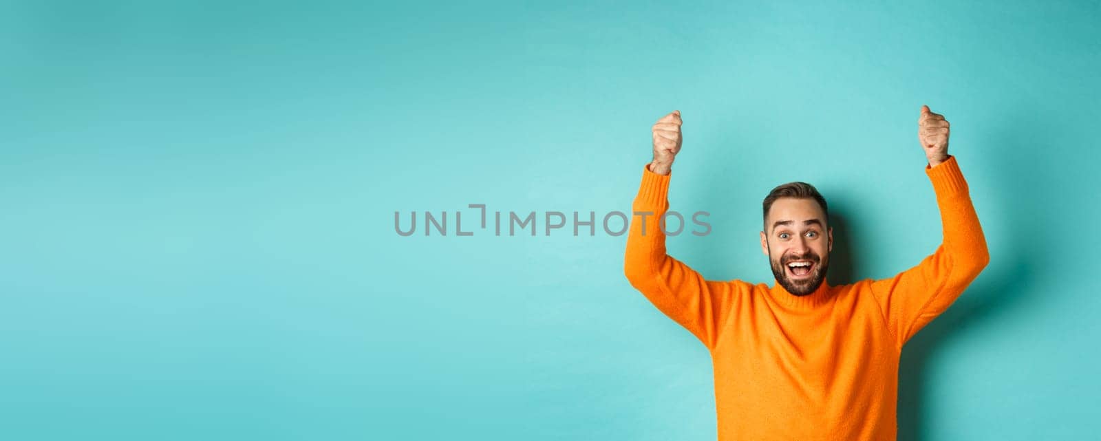 Waist-up shot of happy man raising hands as if holding a sign, showing logo or promo banner, smiling excited, standing in orange sweater against turquoise background.