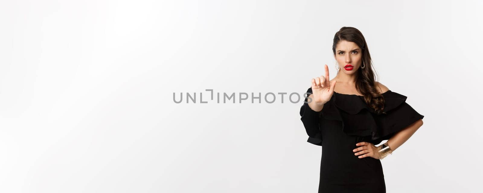 Fashion and beauty. Confident and serious lady in black dress, showing finger in stop gesture, prohibit and disapprove something, standing over white background.