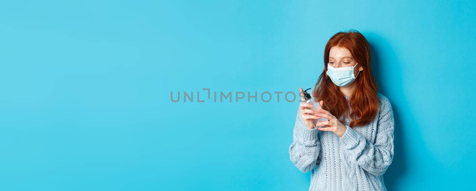 Winter, covid-19 and social distancing concept. Young redhead woman in face mask clean hands with antiseptic, disinfecting with hand sanitizer, standing against blue background by Benzoix