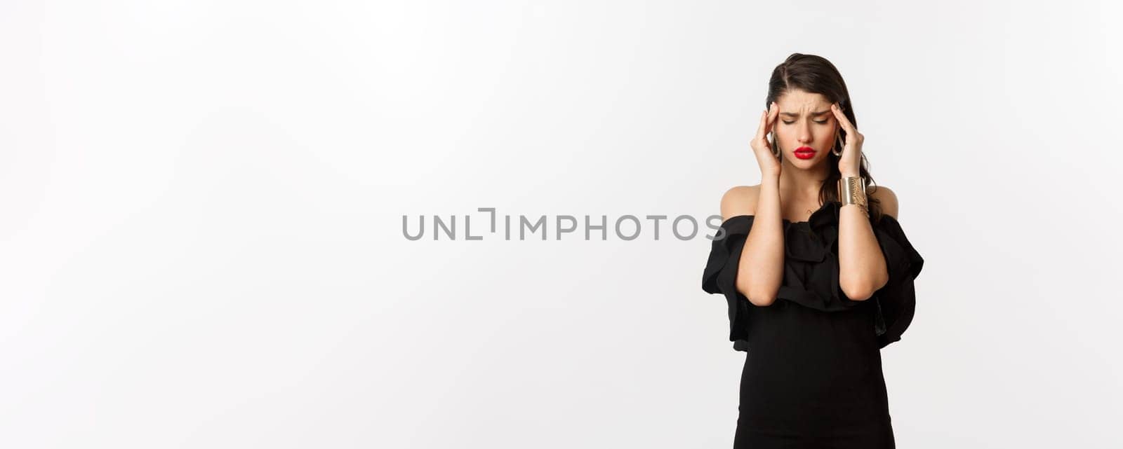 Fashion and beauty. Young modern woman in black dress, red lipstick, having headache, touching head and feeling sick, standing over white background by Benzoix