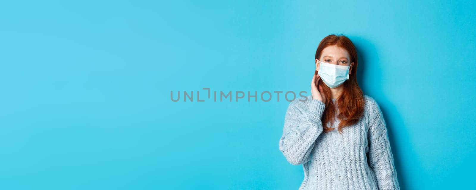 Winter, covid-19 and social distancing concept. Beautiful redhead teenage girl, wearing face mask and tuck hair strand behind ear, staring at camera, standing over blue background by Benzoix