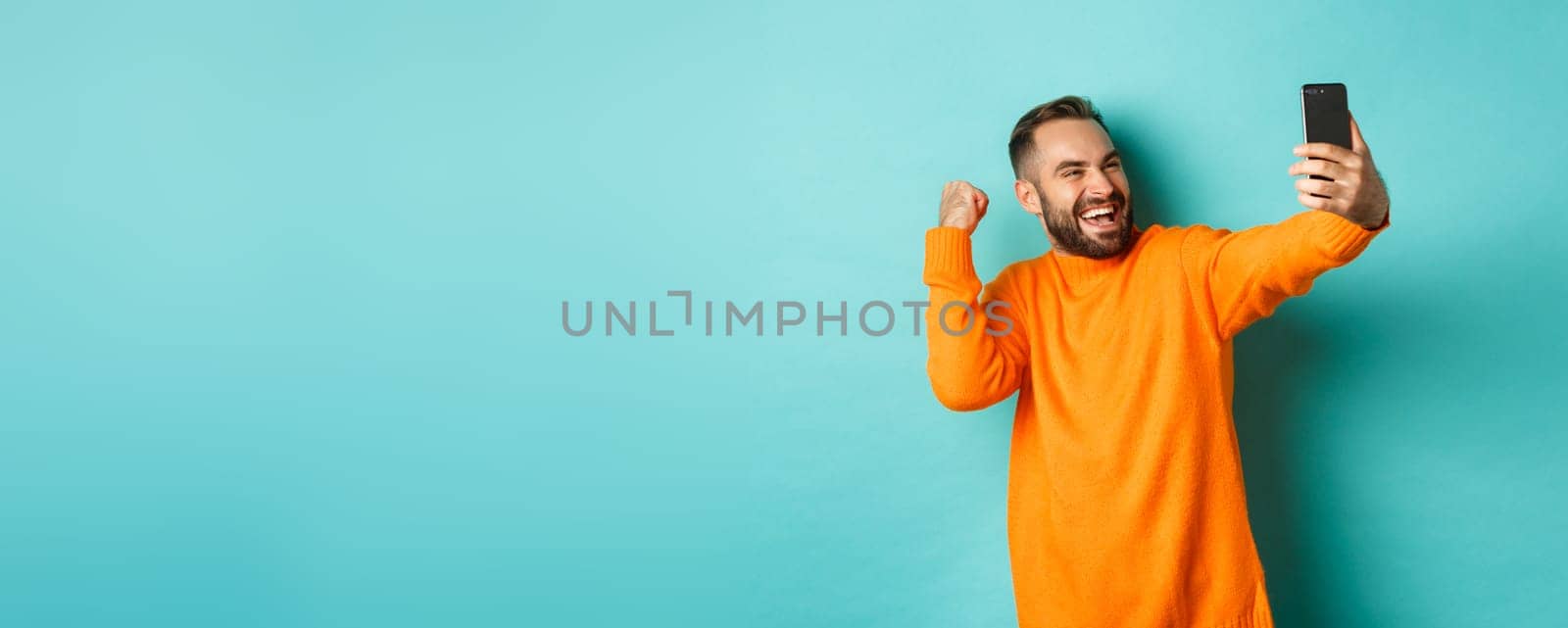 Handsome man video calling, looking happy and rejoicing of good news, making fist pump, communicating via mobile phone, standing over light blue background.