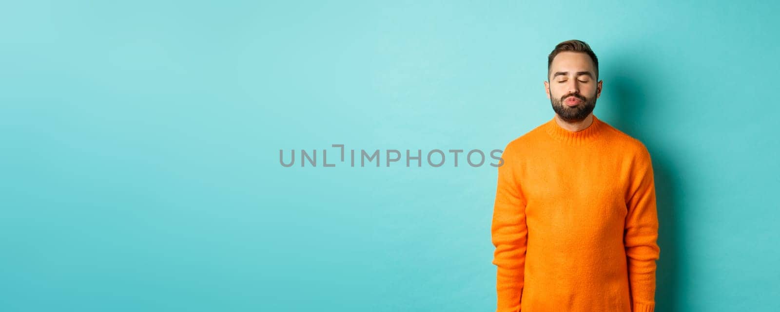 Waist up shot of young man pucker lips and close eyes, waiting for kiss, standing in orange sweater against light blue background by Benzoix