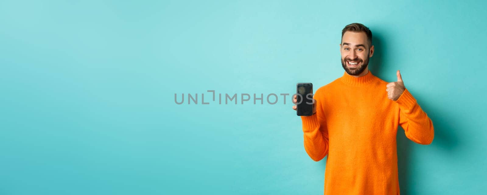 Attractive bearded man showing smartphone screen, thumbs up, recommending mobile app, standing satisfied over turquoise background.