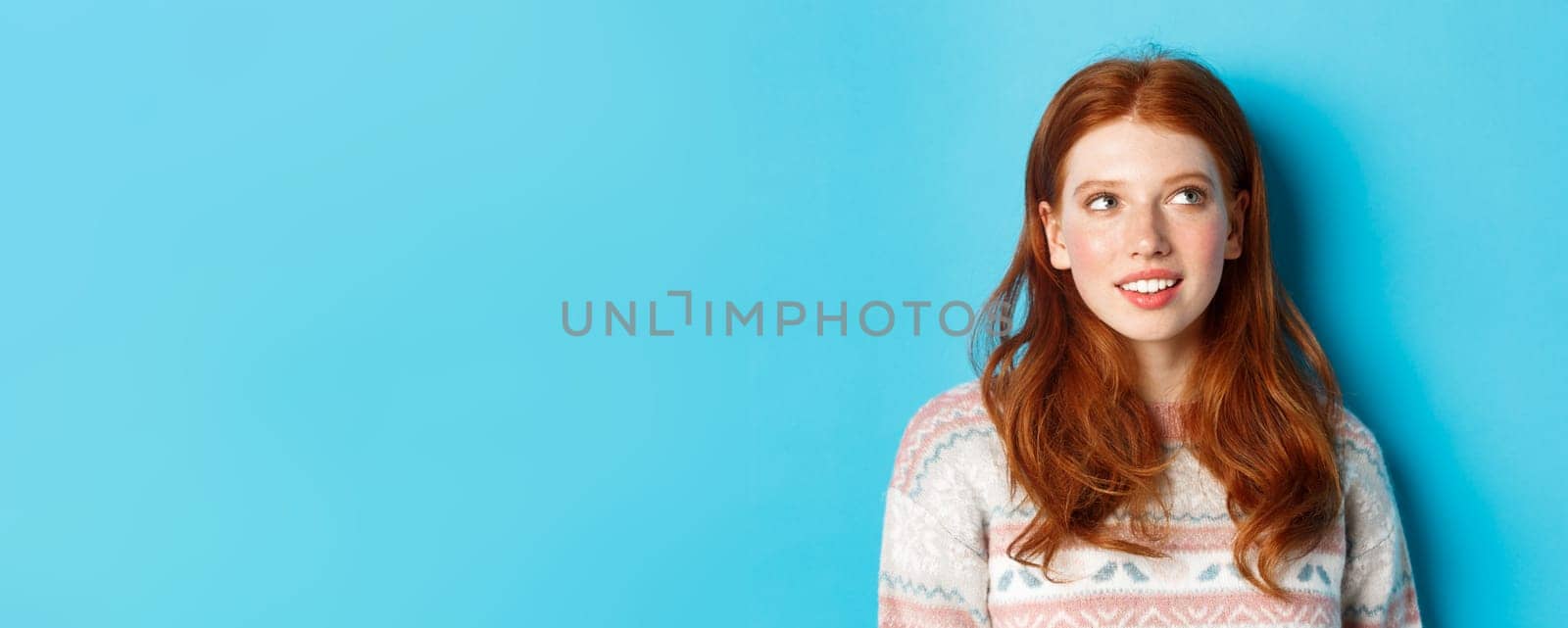 Close-up of dreamy redhead girl imaging something, staring at upper right corner and smiling, standing in winter sweater against blue background by Benzoix