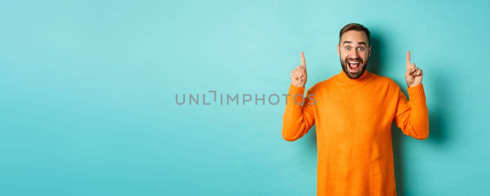 Young happy man showing advertisement, pointing fingers up announcing promo offer, standing over turquoise background.