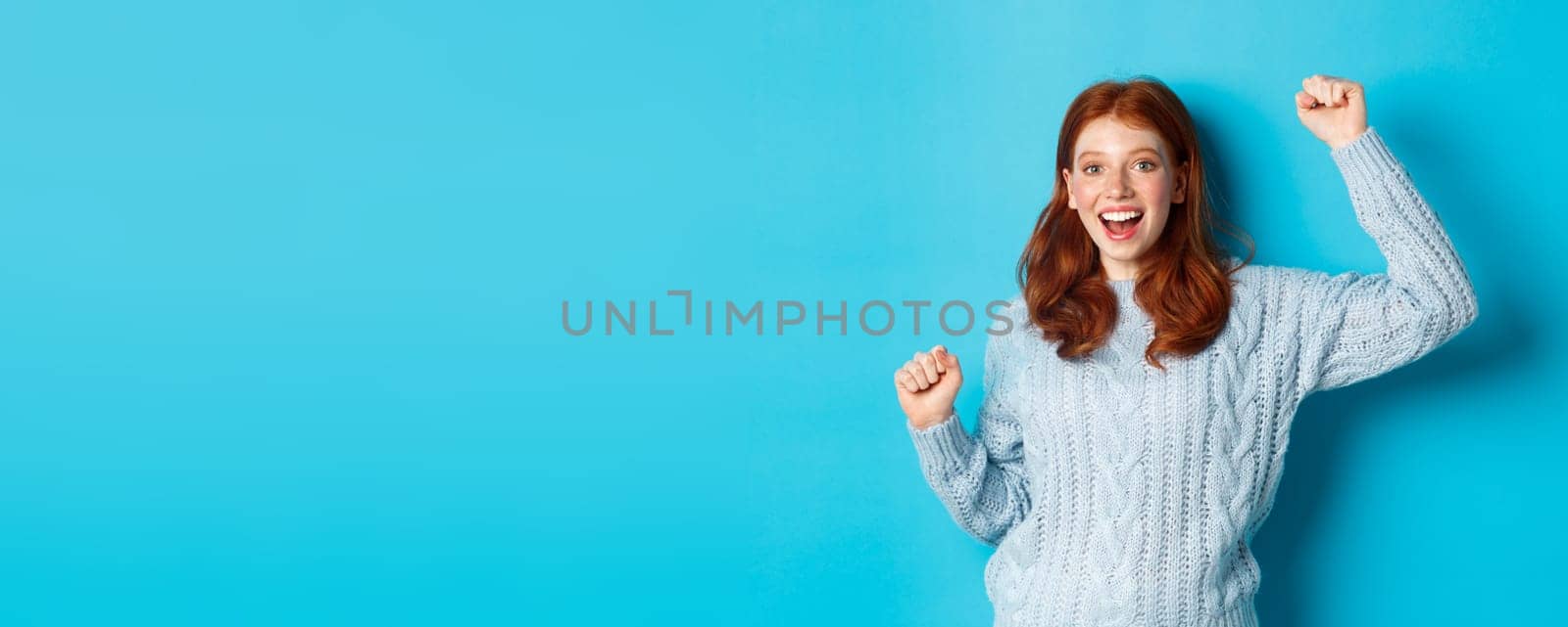 Cheerful redhead gil winning, celebrating victory, smiling and jumping from happiness, posing against blue background.