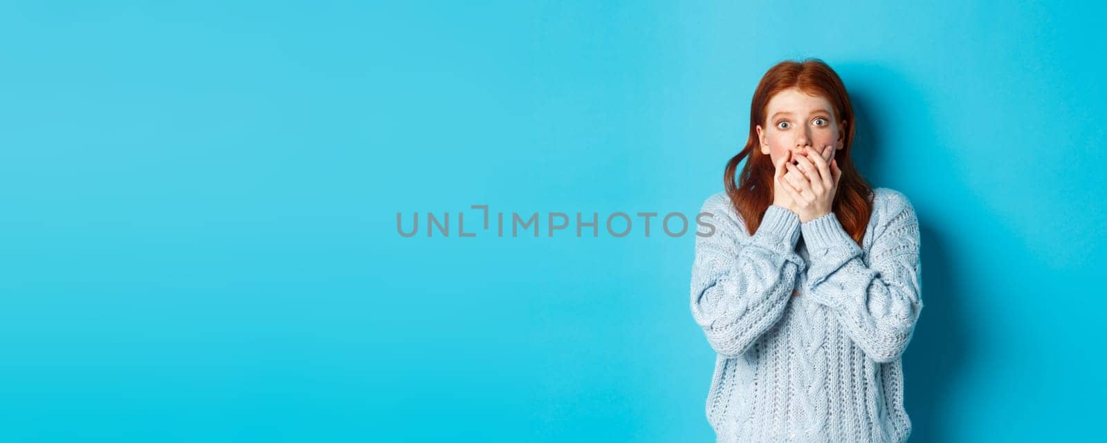Shocked redhead girl in sweater, staring at camera scared and gasping, covering mouth with hands, standing against blue background by Benzoix