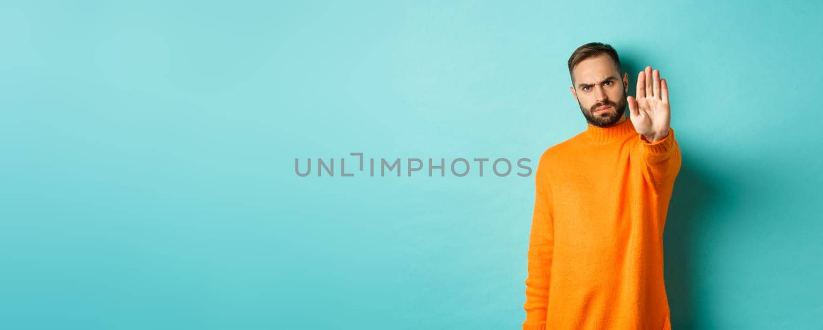 Serious frowning man showing stop gesture, telling no, prohibit and disagree, standing over light blue background.