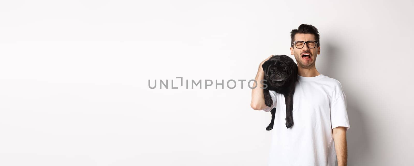 Sad dog owner crying, holding cute black pug on shoulder and looking miserable, sobbing while standing over white background.