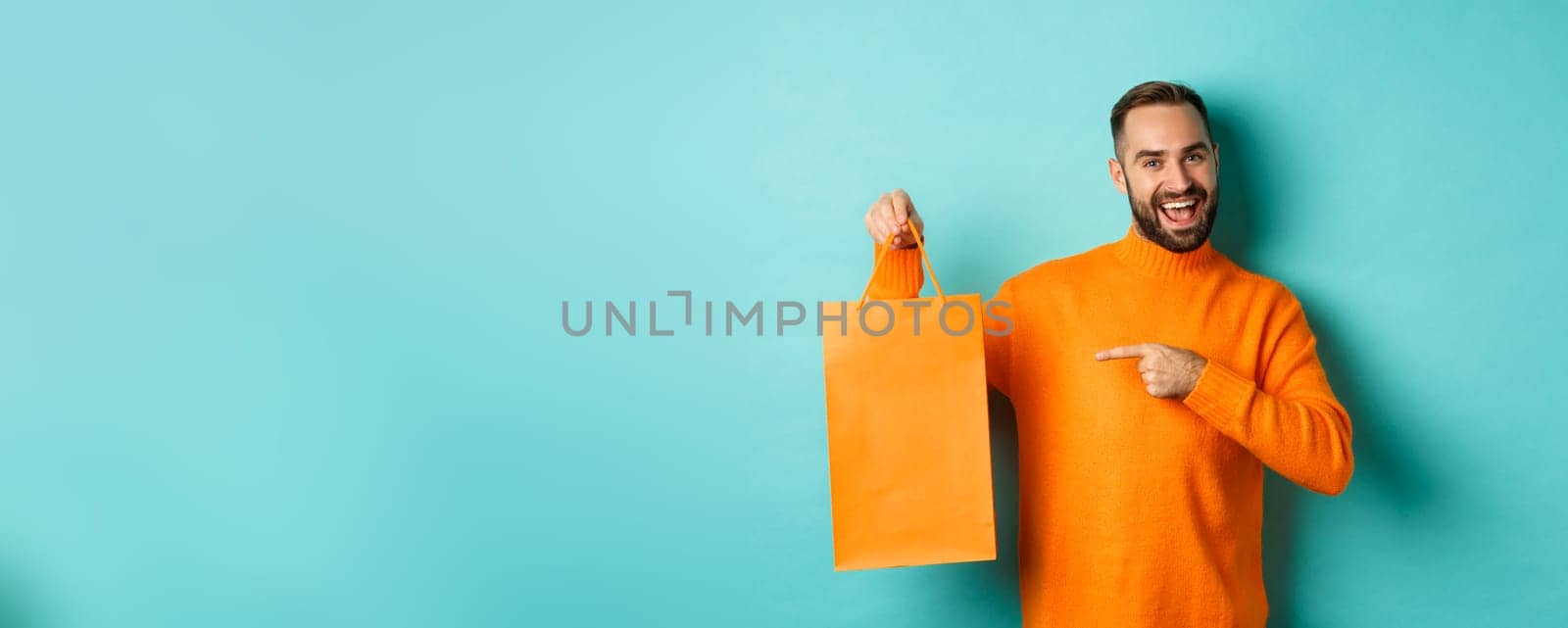Satisfied male customer pointing at orange shopping bag, recommending store, smiling pleased, standing over turquoise background.