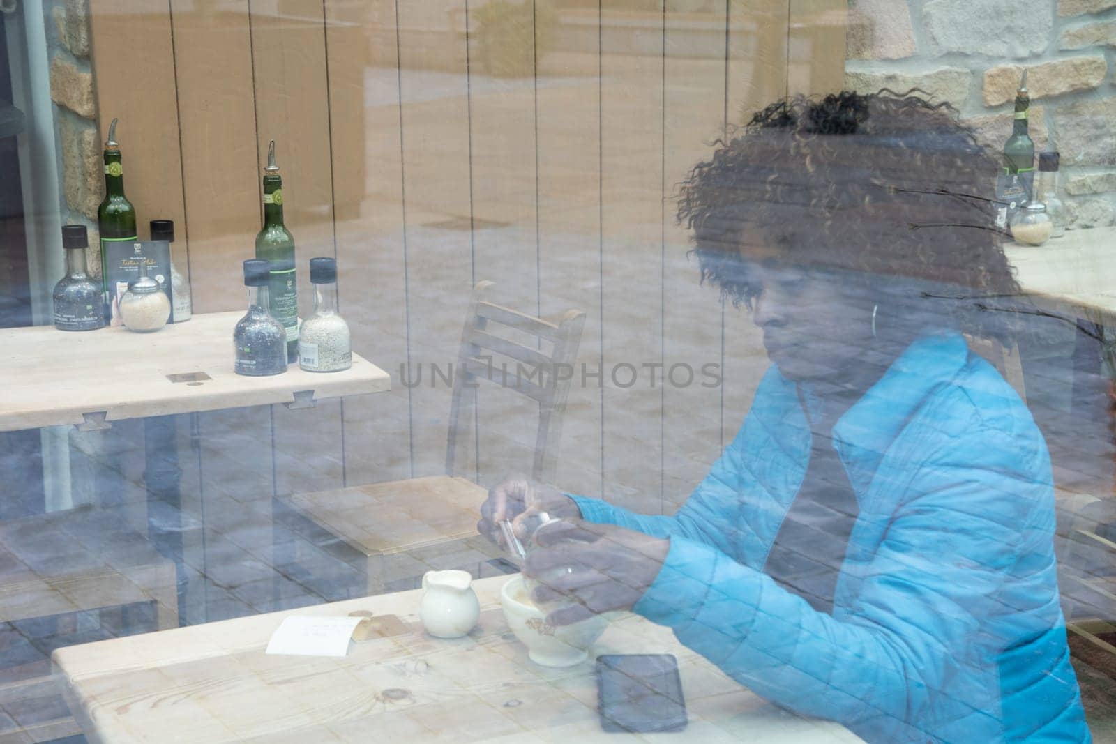 An African American woman in a blue jacket sits behind a window glass in a cafe makes an order using an application on a smartphone, selective soft focus from due to dirty glass. High quality photo