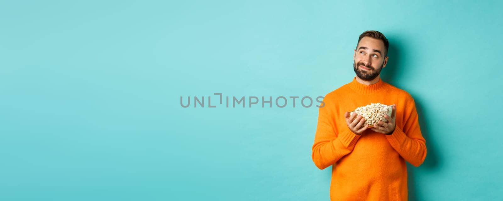 Handsome young man in orange sweater, looking thoughtful at upper left corner, holding popcorn, picking movie, blue background by Benzoix