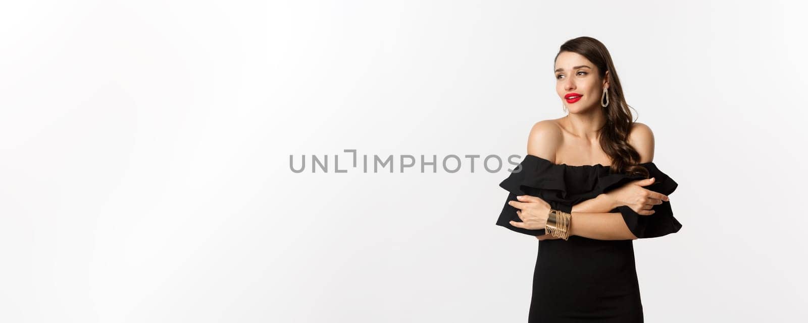 Beauty and fashion concept. Elegant and beautiful woman in black dress, makeup, embracing herself and looking away with sensual gaze, standing over white background by Benzoix