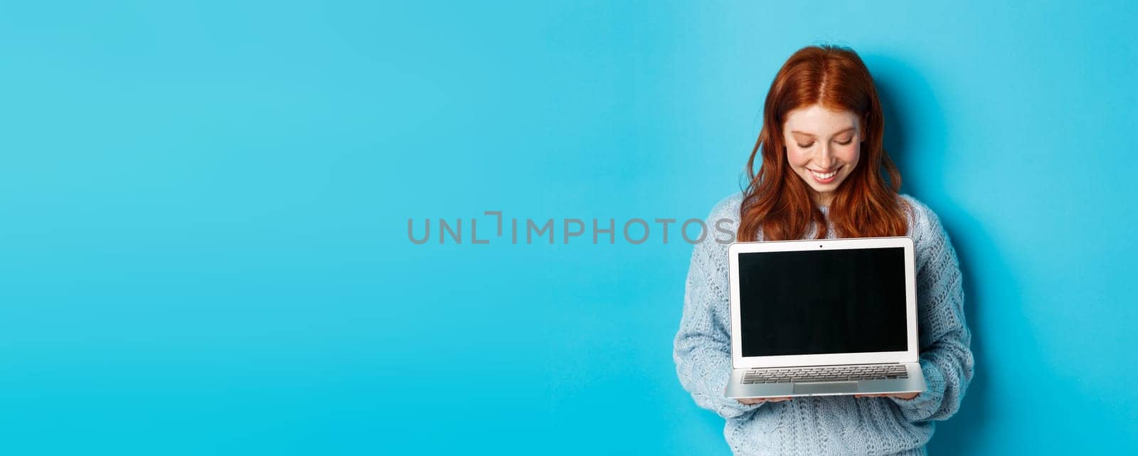 Cute redhead woman in sweater, showing and looking at laptop screen with pleased smile, demonstrating something online, standing over blue background.