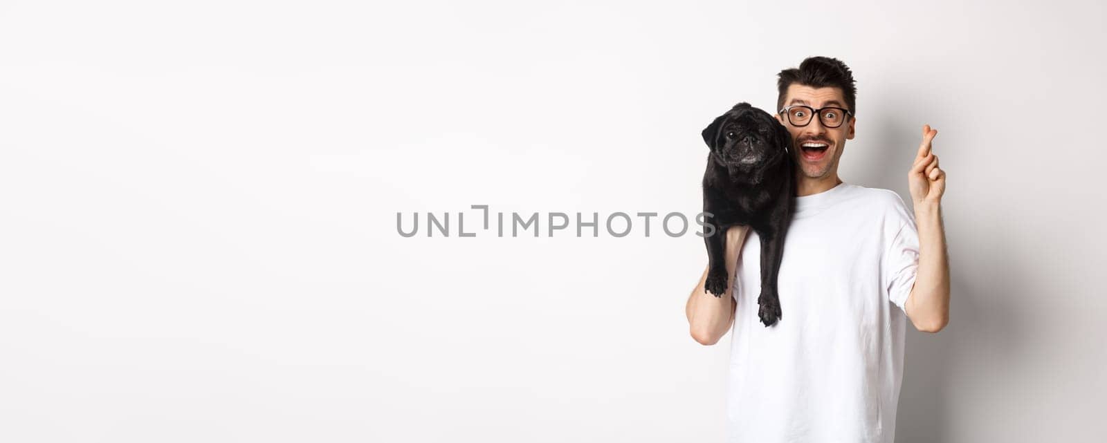 Hopeful smiling dog owner making a wish, holding cute black pug on shoulder and cross fingers for good luck, white background by Benzoix