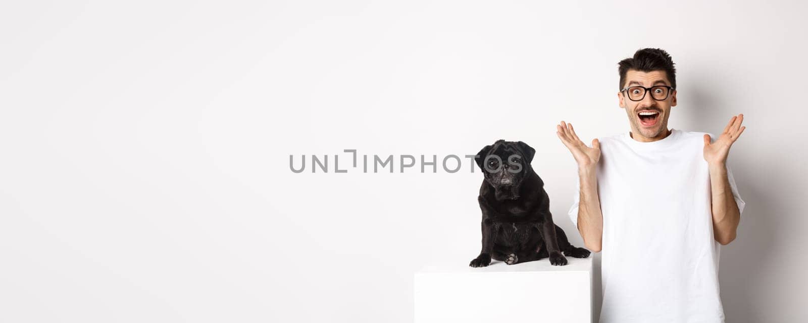 Cheerful young man in glasses standing with his pet, rejoicing and staring at camera amused, hear great news, standing with pug over white background.