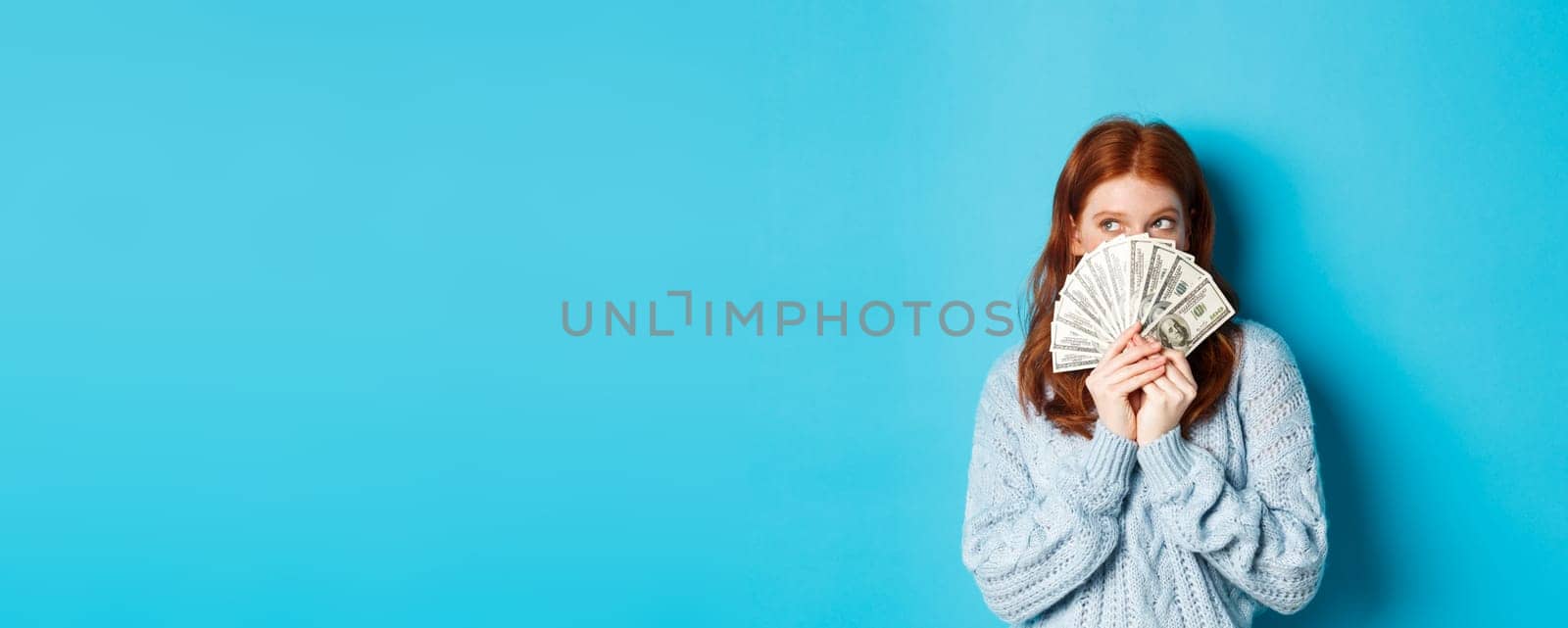 Thoughtful cute girl with red hair dreaming about shopping, holding dollars and looking at upper left corner logo, standing over blue background by Benzoix