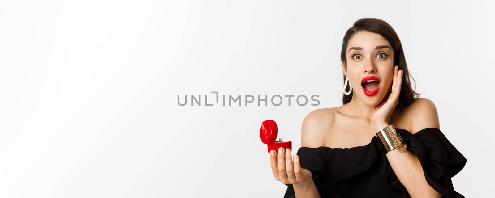 Close-up of attractive young woman with red lipstick, makeup on, looking amazed after receiving marriage proposal, holding engagement ring, standing over white background.