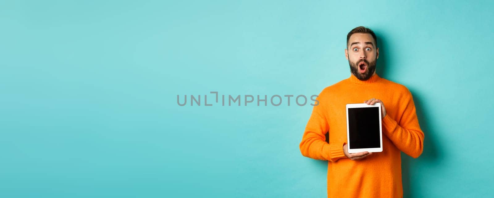 Technology. Surprised bearded man drop jaw, staring at camera and showing digital tablet screen, standing againt light blue background by Benzoix