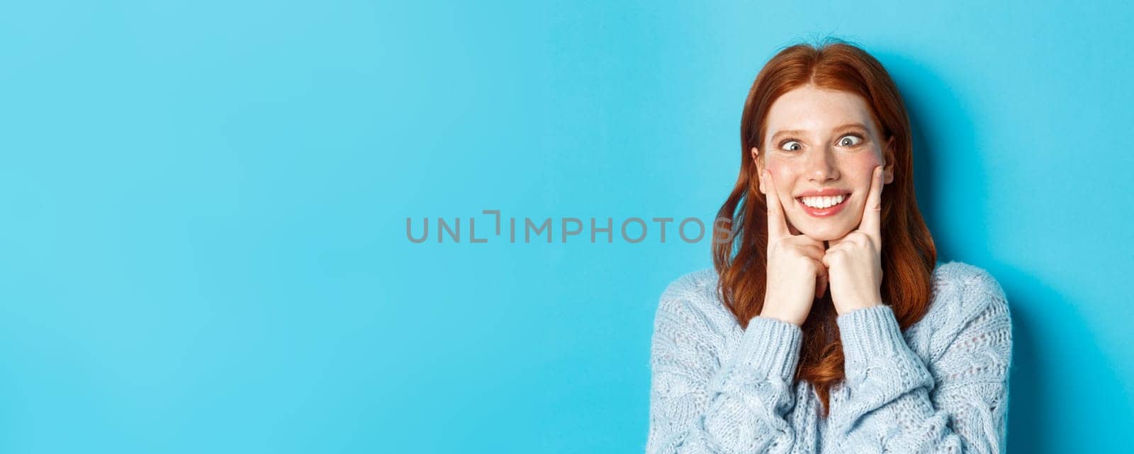 Close-up of funny redhead teen girl making faces, squinting and squeezing cheeks, standing against blue background.