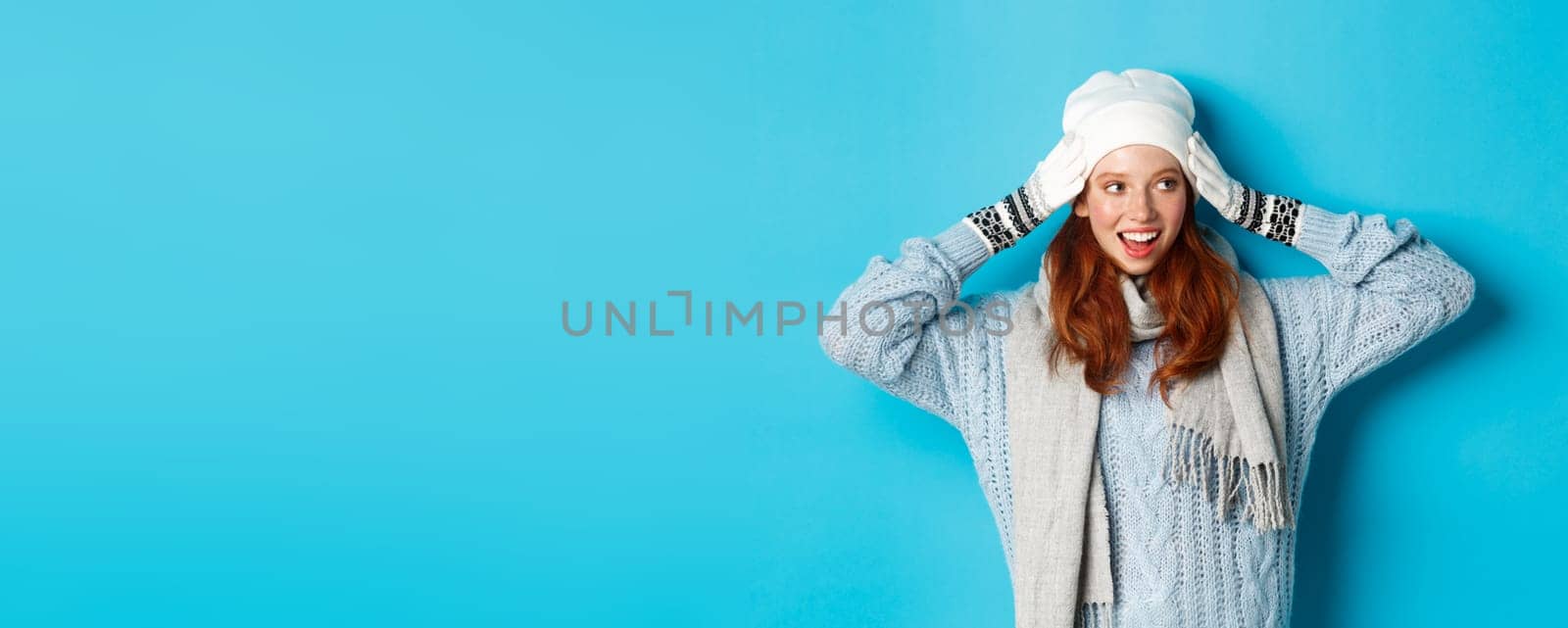 Winter and holidays concept. Happy redhead girl in beanie hat, scarf and gloves looking right and smiling, standing against blue background.