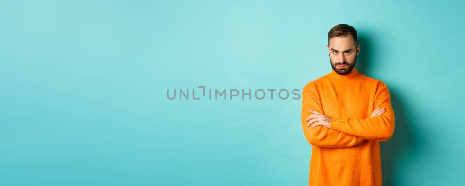 Offended man looking angry at you, cross arms on chest and stare mad, standing in orange sweater against turquoise background by Benzoix