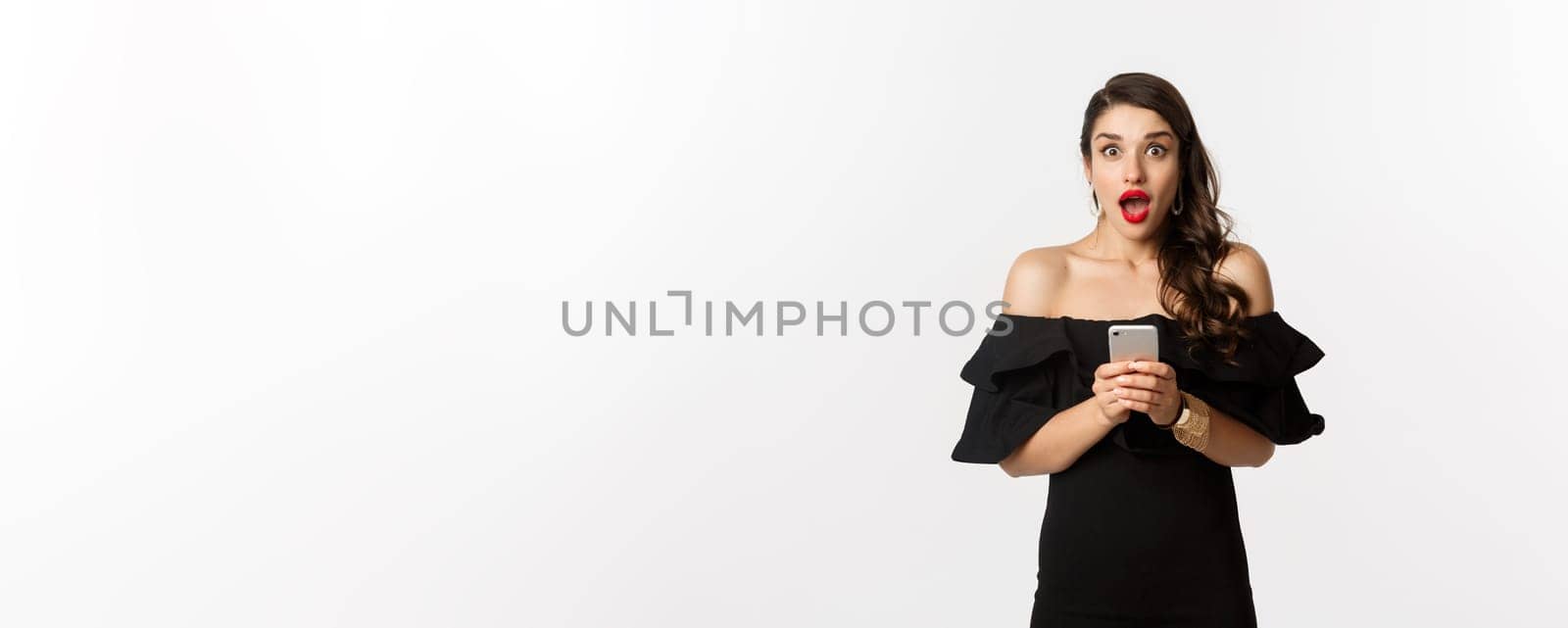 Happy young glamour woman in black dress, holding mobile phone and looking surprised, standing over white background.