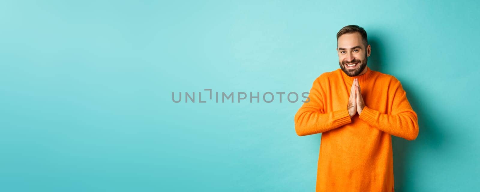 Happy man saying thank you, holding hands in pray, looking grateful and smiling, standing over light blue background.