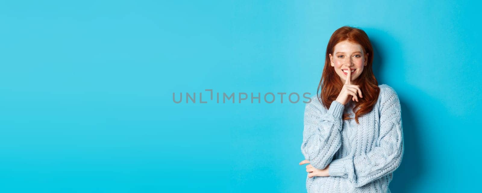 Pretty redhead teenager hushing and smiling, telling a secret, standing in sweater against blue background by Benzoix