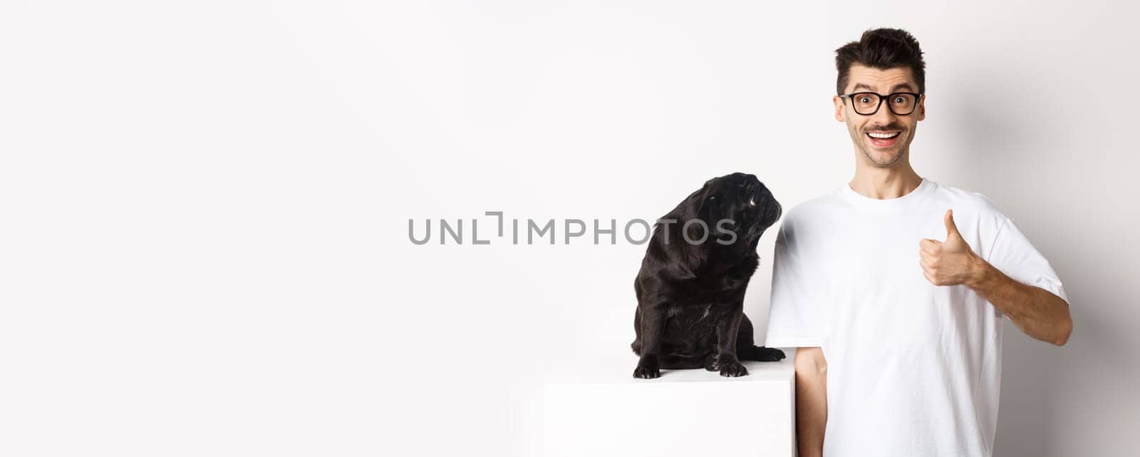 Cheerful handsome guy standing near cute black pug and showing thumb-up. Pet owner approve and recommend product for dogs, white background by Benzoix
